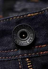 Musta metallinen painike, jossa on teksturoitu pinta ja teksti "SUPERDRY STANDARD BLUE JEANS", tummansinisen denimmateriaalin päällä oransseilla saumoilla.