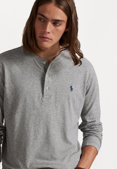 Γκρι πουκάμισο henley με μακριά μανίκια, κατασκευασμένο από απαλό βαμβάκι, διαθέτοντας τρεις κουμπότρυπες στο λαιμό και ένα μικρό ναυτικό λογότυπο στο αριστερό στήθος.