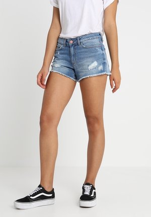 Vrouw draagt lichtblauwe versleten spijkershorts, wit T-shirt en zwart-witte sneakers, staand tegen een effen witte achtergrond.