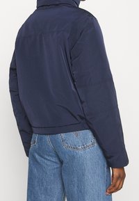 Person klädd i en marinblå jacka och ljusblå denimjeans, stående med ryggen mot kameran framför en enfärgad bakgrund.