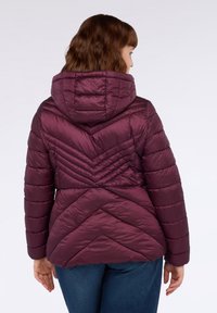 Kvinna som bär en huvad, vadderad maroon pufferjacka sedd bakifrån, tillsammans med blå jeans mot en enkel ljus bakgrund.