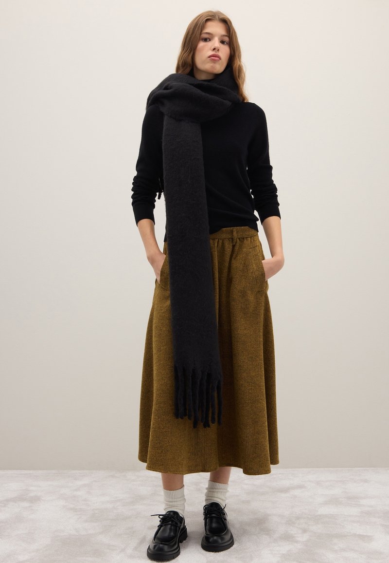 Sciarpa di lana nera avvolta intorno al collo; abbinata a una maglia nera a maniche lunghe e a una gonna color senape lunga fino al ginocchio; scarpe nere con suole spesse.