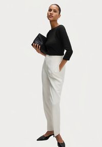 Schwarzes Langarmshirt, weiße hoch taillierte, maßgeschneiderte Hose und schwarze spitze Ballerinas; hält eine schwarze, gewebte Clutch-Tasche.