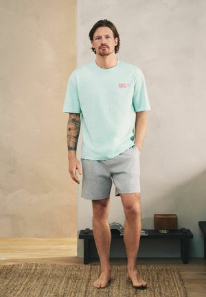 Mand med tatoveret arm står barfodet på et tæppe iført lyseblå "Daily Grind" T-shirt og grå shorts i et minimalistisk rum med lav hylde.