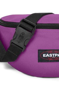 Violetti kangaskassi mustalla hihnalla, jossa on vetoketjutasku ja muovinen solki. Kassi esittelee Eastpak-logon mustalla tarralla.