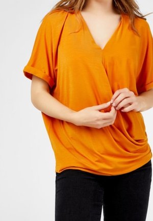 Donna che indossa una maglia arancione ampia stile incrociato con maniche corte arrotolate e pantaloni neri, mani delicatamente intrecciate davanti.