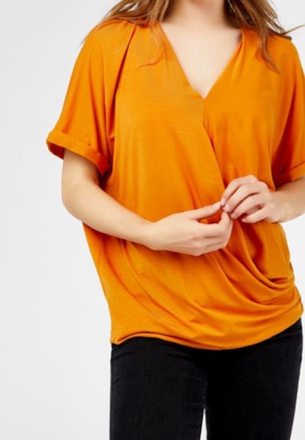 Femme portant un haut orange ample style cache-cœur avec manches courtes retroussées et pantalon noir, les mains délicatement jointes devant.