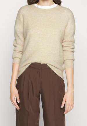 Femme portant un pull beige à manches longues en maille par-dessus une chemise blanche et un pantalon ample marron taille haute, sur un fond uni.