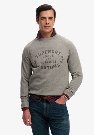 Mężczyzna ubrany w szarą bluzę Superdry na czerwoną kratowaną koszulę, niebieskie dżinsy i zegarek na nadgarstku, stoi z jedną ręką w kieszeni.