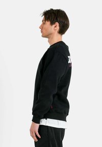 Zwarte katoenen sweatshirt met tekst op de rug; heeft een ronde hals en lange mouwen; gedragen over een witte gelaagde shirt.