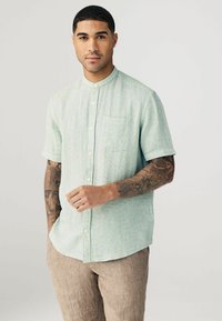 Camisa de botón de manga corta color verde claro, con tejido texturizado, que presenta un pequeño bolsillo en el pecho. Combinada con pantalones de lino beige, mostrando tatuajes en el brazo.