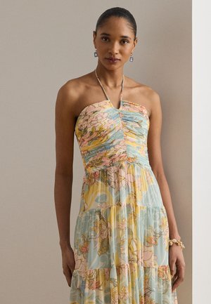 Γυναίκα με μαλλιά τραβηγμένα προς τα πίσω, που φοράει ένα παστέλ φόρεμα με λουλούδια και σκουλαρίκια με μπλε πολύτιμους λίθους, στέκεται μπροστά σε έναν ουδέτερο τοίχο.