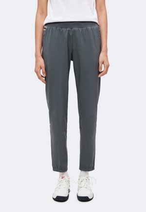Persona con pantalones de cintura elástica de color gris oscuro y zapatillas blancas con calcetines blancos, de pie contra un fondo claro y liso.