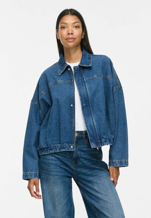Mujer con cabello largo y oscuro que lleva una chaqueta vaquera azul sobre una camiseta blanca y vaqueros azules, de pie frente a un fondo blanco liso.