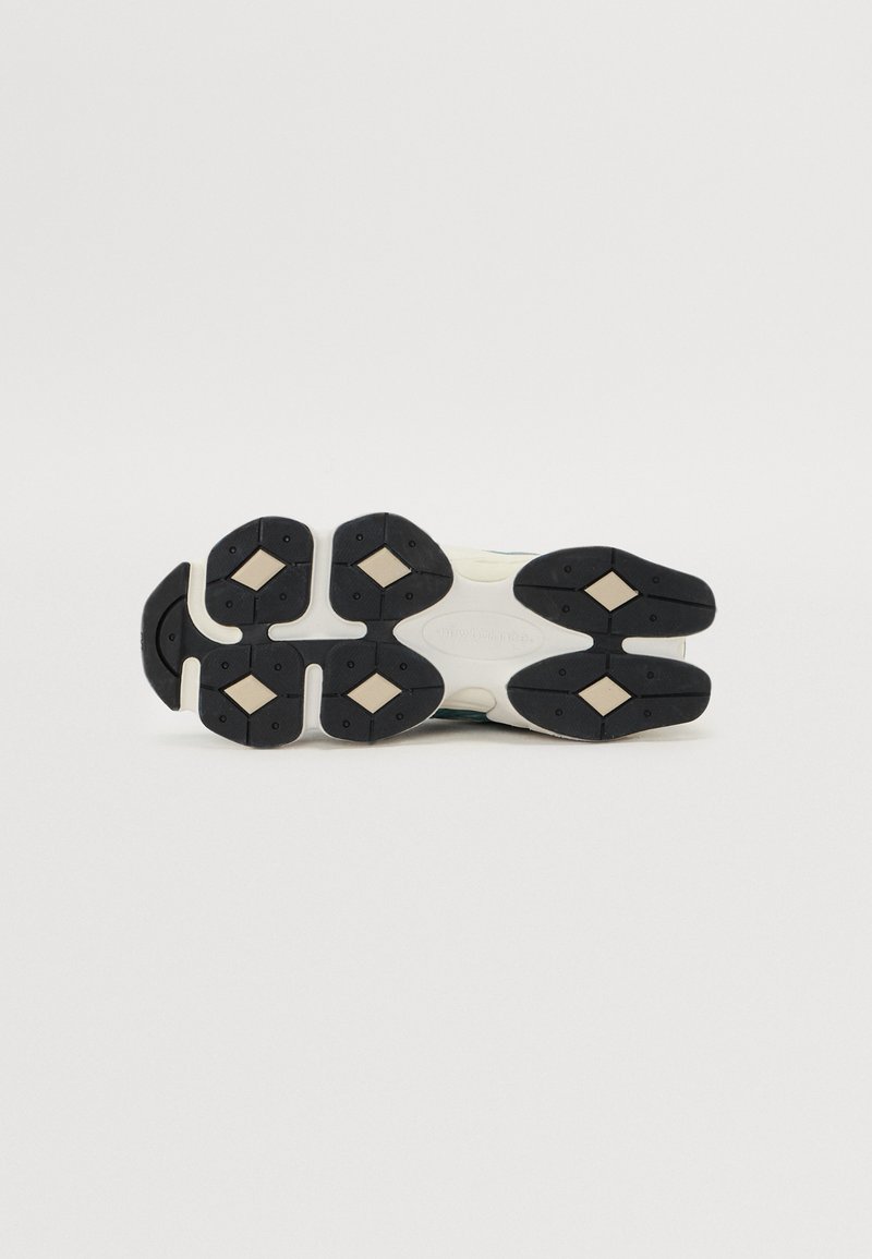 Schwarze Gummi-Außensohle mit Diamantmustern, cremefarbene Zwischensohle und grüne sowie blaue Akzente im Obermaterial. Runde Logodetail vorhanden.