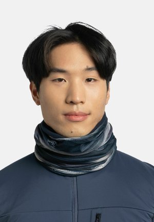 Joven con pelo lacio negro que lleva una chaqueta azul oscura y un cuello de botella con un patrón en negro y gris.