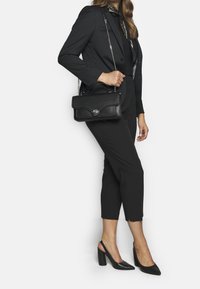 Černá kožená kabelka s texturovaným povrchem, s klopou a stříbrnými detaily. Modelka má na sobě elegantní černé kalhoty a podpatky.
