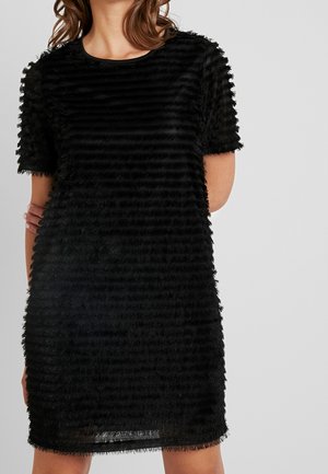 Mujer vestida con un vestido negro de manga corta y textura, con franjas horizontales con flecos, de pie sobre un fondo claro y simple.