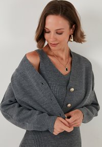 Vestido de punto gris con un cárdigan gris abierto que presenta grandes botones dorados. Textura suave con silueta ajustada y aspecto en capas.