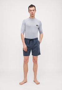 Szürke rövid ujjú fürdőfelső Quiksilver logóval, sötétkék fürdőnadrággal párosítva, amely rugalmas derékkal és zsinórral rendelkezik. Mezítláb álló modell.