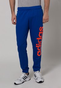 Modré tepláky s elastickým páskem, na levé noze s oranžovým logem "adidas". Vyrobené z měkké, strukturované tkaniny.