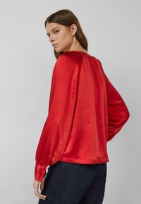 Frau mit braunen Haaren, die eine locker sitzende rote Satinbluse und dunkle Hosen trägt, posiert mit dem Rücken zur Kamera und leicht seitlich gedrehtem Kopf.