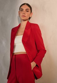 Rød skræddersyet jakkesætjakke med noterede slag, parret med en hvid crop top. Glat tekstur, struktureret form, diskret skulderpolstring og lommer.