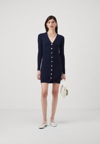 Marineblauw geribbeld jurkje met lange mouwen, V-hals en knopen aan de voorkant. In combinatie met een witte handtas en instappers. Glad textuur.