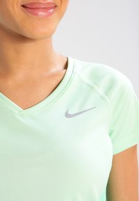 Persona sonriente vistiendo una camiseta deportiva de Nike de color verde claro con cuello en V y un logo de Nike en color gris visible en el pecho.