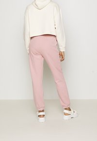 Person som bär ljusrosa joggers, vit kort hoodie och vita sandaler med pärlprydnader, stående mot en enfärgad bakgrund.