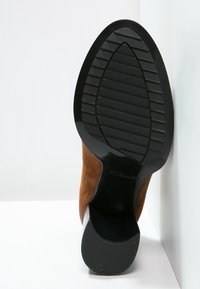 Zapato de tacón alto de ante marrón con suela de goma negra, que presenta líneas de agarre texturizadas y acentos lisos en los laterales.