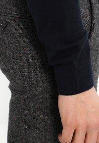 Pantaloni di costume in tweed grigio con fili multicolori. Parte del polsino a maglia di un maglione nero è parzialmente visibile sul lato sinistro. Texture liscia.