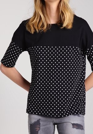 Woman wearing a black short-sleeve top with white polka dots and distressed light gray jeans, standing with hands behind her back.