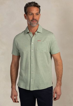 Man draagt een lichtgroen overhemd met korte mouwen, een borstzakje en zwarte broek, staande tegen een effen achtergrond.