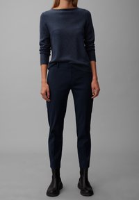 Kobieta w granatowym swetrze dzianinowym, dopasowanych granatowych spodniach i czarnych botkach za kostkę, stojąca na jednolitym szarym tle.