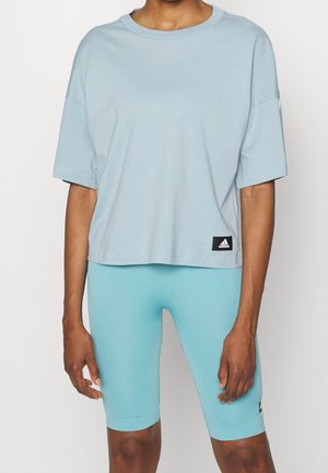 Persona che indossa una maglietta larga azzurro chiaro e pantaloncini da ciclismo aderenti fino al ginocchio dello stesso colore, su uno sfondo uniforme.