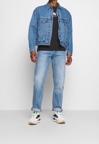 Denim jacket with two chest pockets, light blue jeans, and white sneakers featuring black and red accents and green laces. Black t-shirt underneath.
