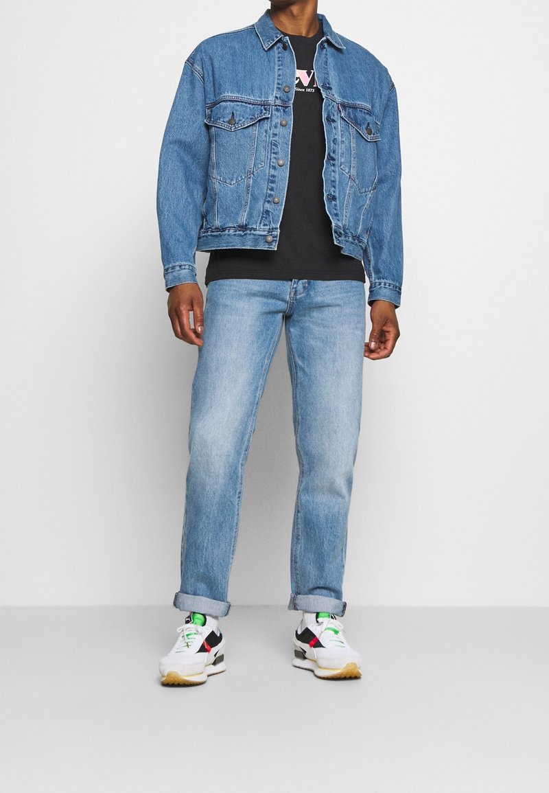Denim jacket with two chest pockets, light blue jeans, and white sneakers featuring black and red accents and green laces. Black t-shirt underneath.