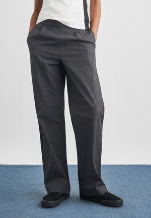 Persona con pantaloni grigi a zampa d'elefante a quadri, scarpe da ginnastica nere e top bianco, in piedi su un tappeto blu con le mani nelle tasche.