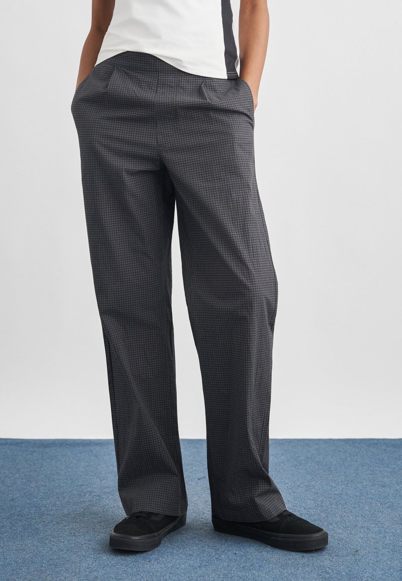 Άτομο που φορά γκρι καρό παντελόνι φαρδιάς γραμμής, μαύρα αθλητικά παπούτσια και λευκό μπλουζάκι, στέκεται πάνω σε μπλε χαλί με τα χέρια στις τσέπες.