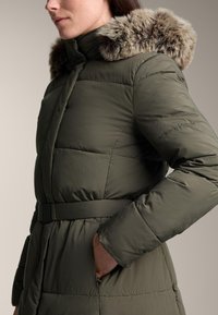 Zateplený olivovo zelený kabát s opaskom v páse a kapucňou s umelou kožušinou. Má prešívanú textúru a bočné vrecká.