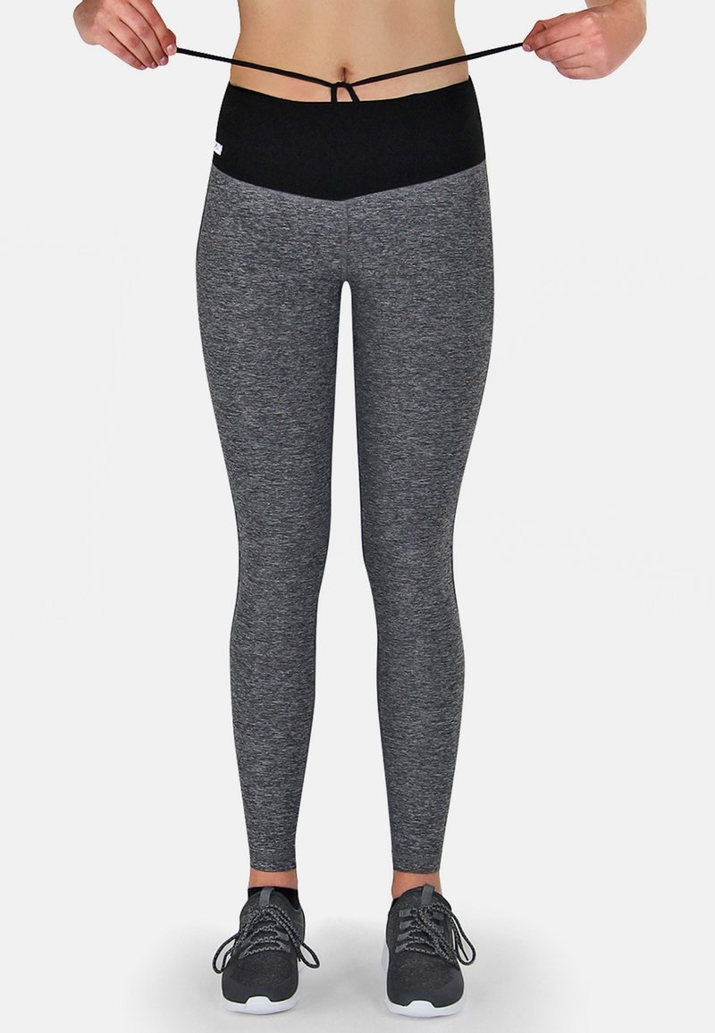 Graue Leggings mit einem hohen schwarzen Bund, der eine Zugkordel hat. Der Stoff wirkt dehnbar und glatt. Schwarze Sportschuhe runden das Outfit ab.