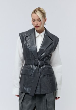Blonde woman wearing a black belted faux leather vest over a white shirt and gray pinstriped pants, looking downward against a plain background.