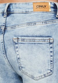 Vaqueros de mezclilla azul claro con un bolsillo trasero y etiqueta de cuero marrón. Presenta un lavado desgastado y detalles de costura visibles.