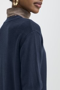 Közeli kép egy személyről, aki sötétkék pulóvert visel bézs színű garbó fölött, arany karika fülbevalókkal, egy egyszerű háttér előtt.