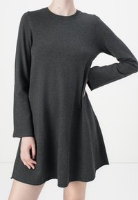 Femme portant une robe A-line à manches longues de couleur gris foncé, se tenant avec une main sur la hanche et l'autre détendue à ses côtés.