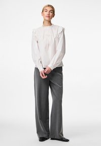 Blouse blanche avec un col et des manches à volants, accompagnée d'un pantalon large gris. La blouse présente une coupe décontractée et une texture en coton.
