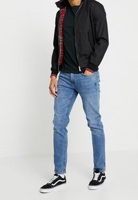 Man som bär en svart bomberjacka med röd rutig foder, svart t-shirt, ljusblå slim-jeans och svarta låga sneakers med vita snörning.