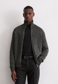 Donkergroen, geribbelde ritssluitingsjack met een hoge kraag, passende mouwen, gedragen over een zwart shirt, in combinatie met zwarte broek en een riem.