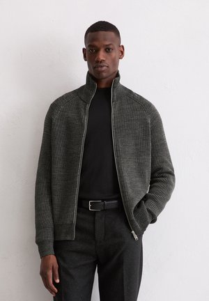 Donkergroen, geribbelde ritssluitingsjack met een hoge kraag, passende mouwen, gedragen over een zwart shirt, in combinatie met zwarte broek en een riem.